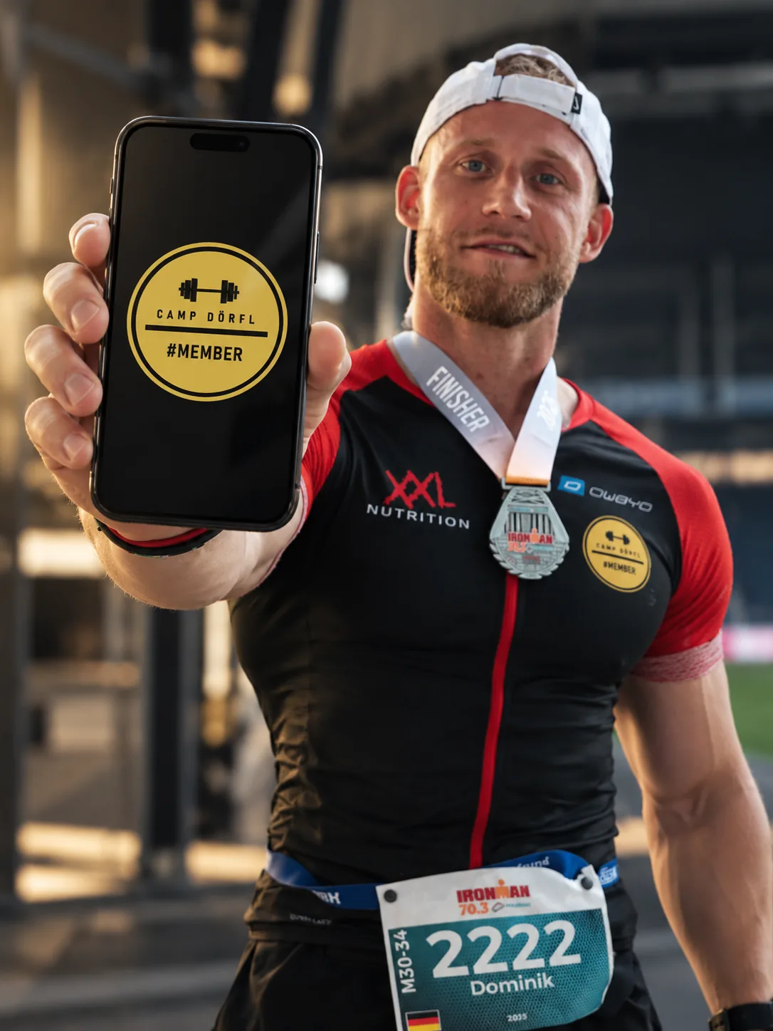 Dominik Dörfl als Ironman-70.3-Finisher hält ein Smartphone mit dem Camp-Dörfl-Member-Zeichen in die Kamera